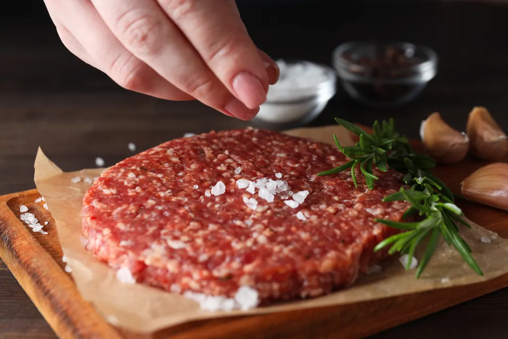 Raw burger patty being seasoned with salt and pepper